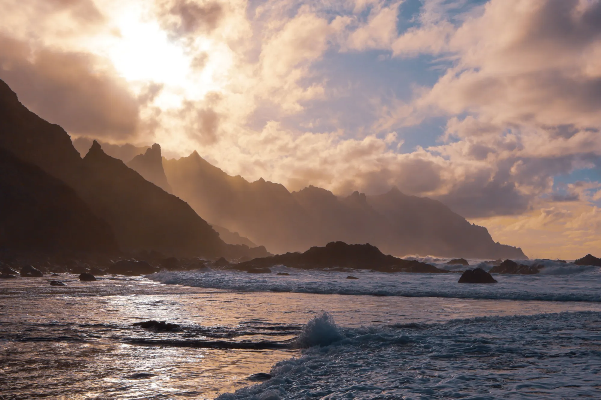 Sunlight breaking through clouds over the jagged Anaga coast, with waves on a dark beach
