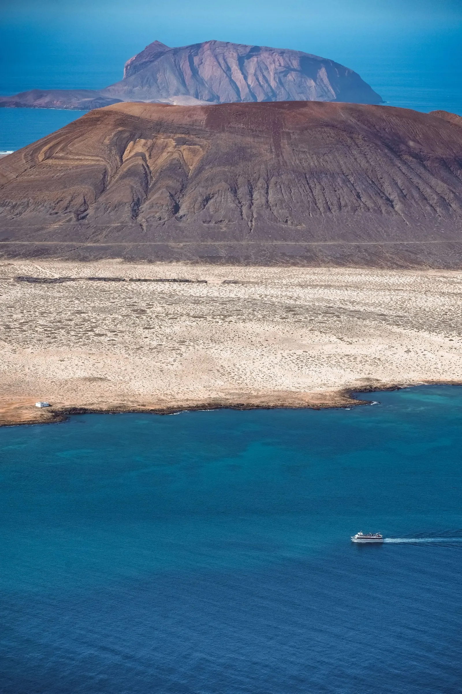 A small ferry crossing the deep blue strait towards La Graciosa, with a brown volcano and Montaña Clara behind