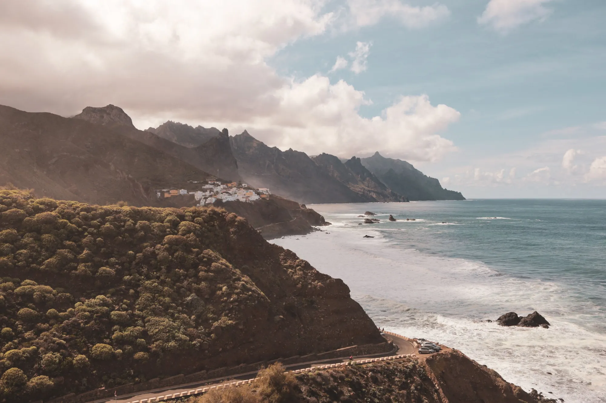 Jagged mountains running down to the sea, with a white village on a ridge and a coast road below