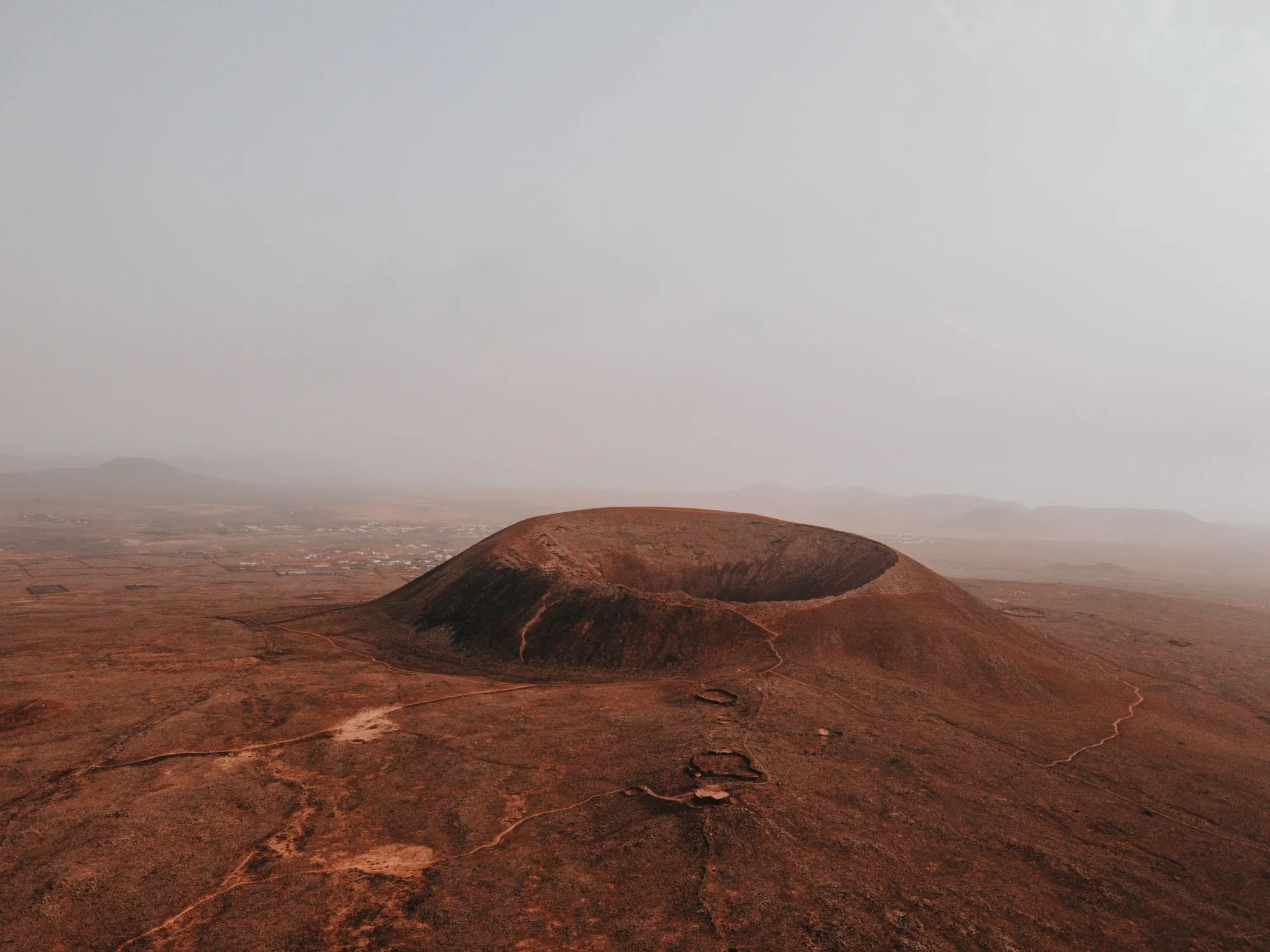 A round volcanic crater on a red-brown plain, with a footpath leading up to the rim