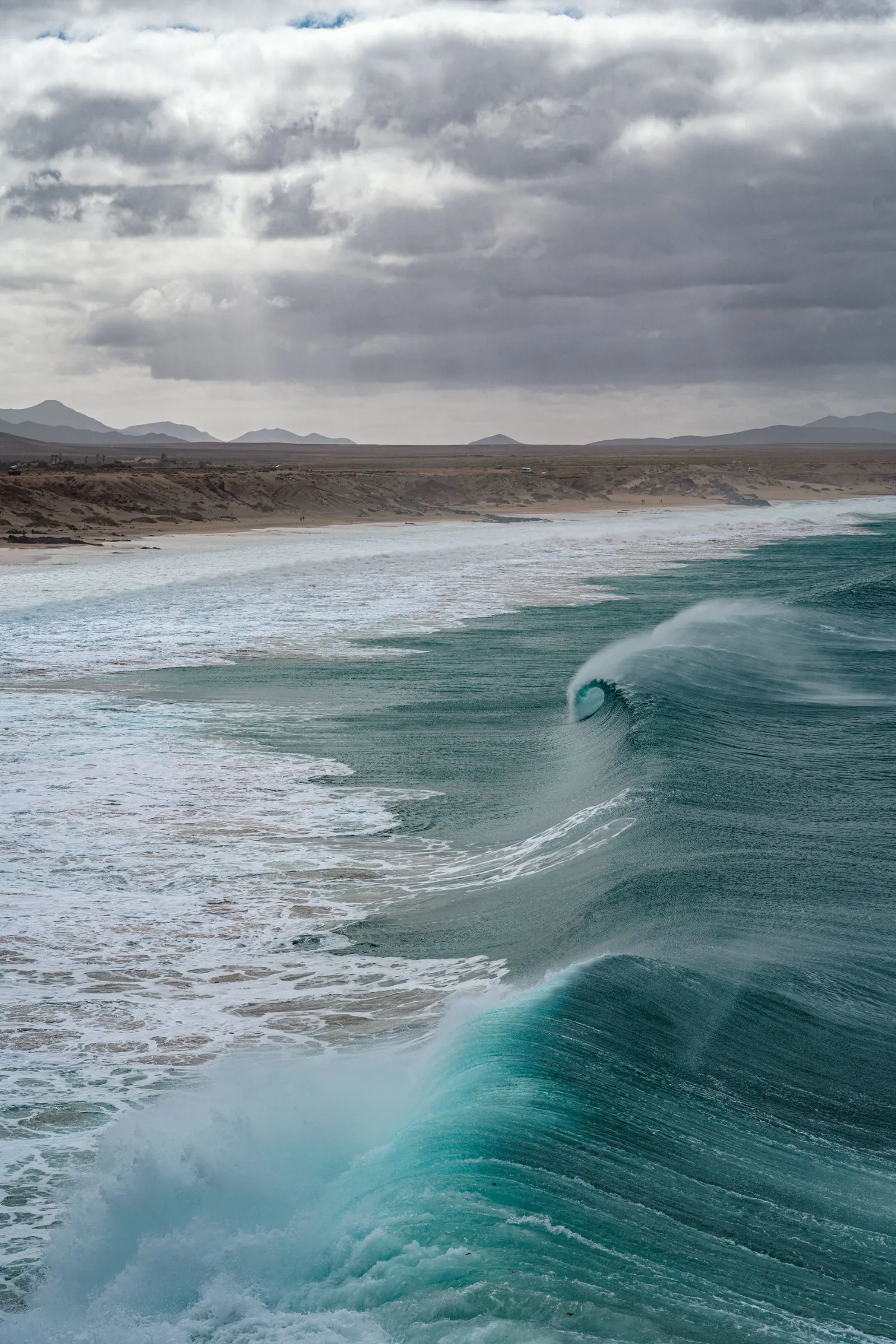 A turquoise wave curling towards a long sandy beach under grey clouds