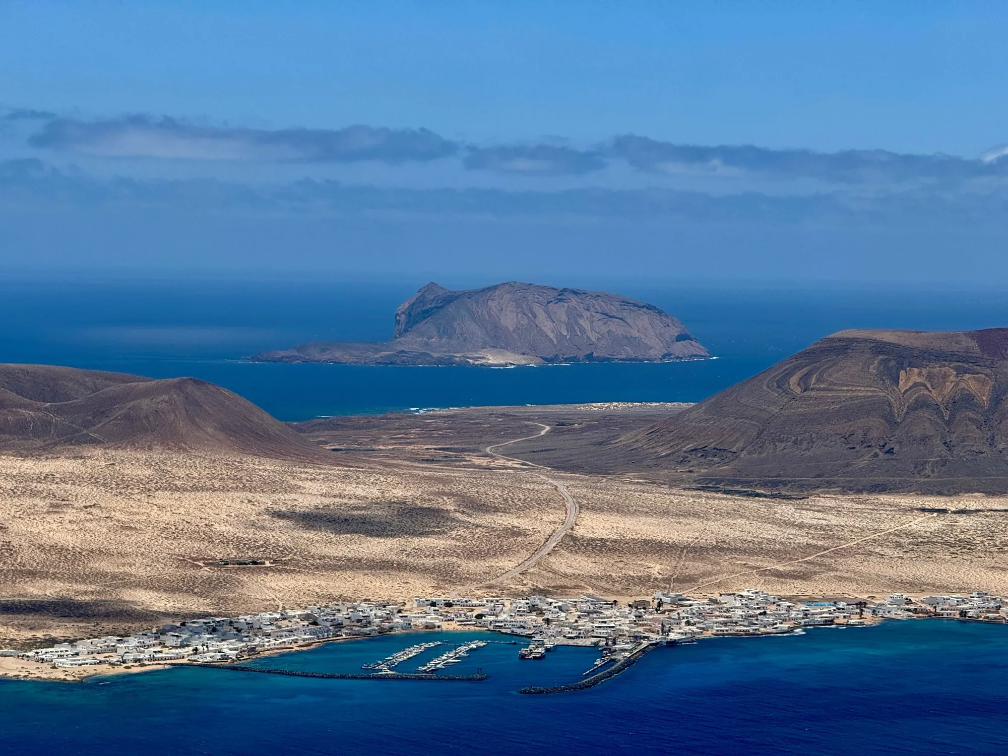 White houses and a small harbour below a sandy plain and volcanoes, with Montaña Clara in the sea behind