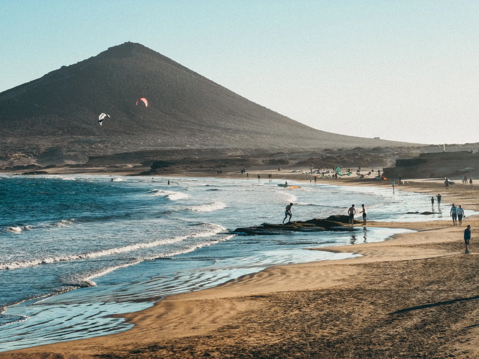 Kitesurfers on the sea in front of a cone-shaped mountain, with people walking on a sandy beach